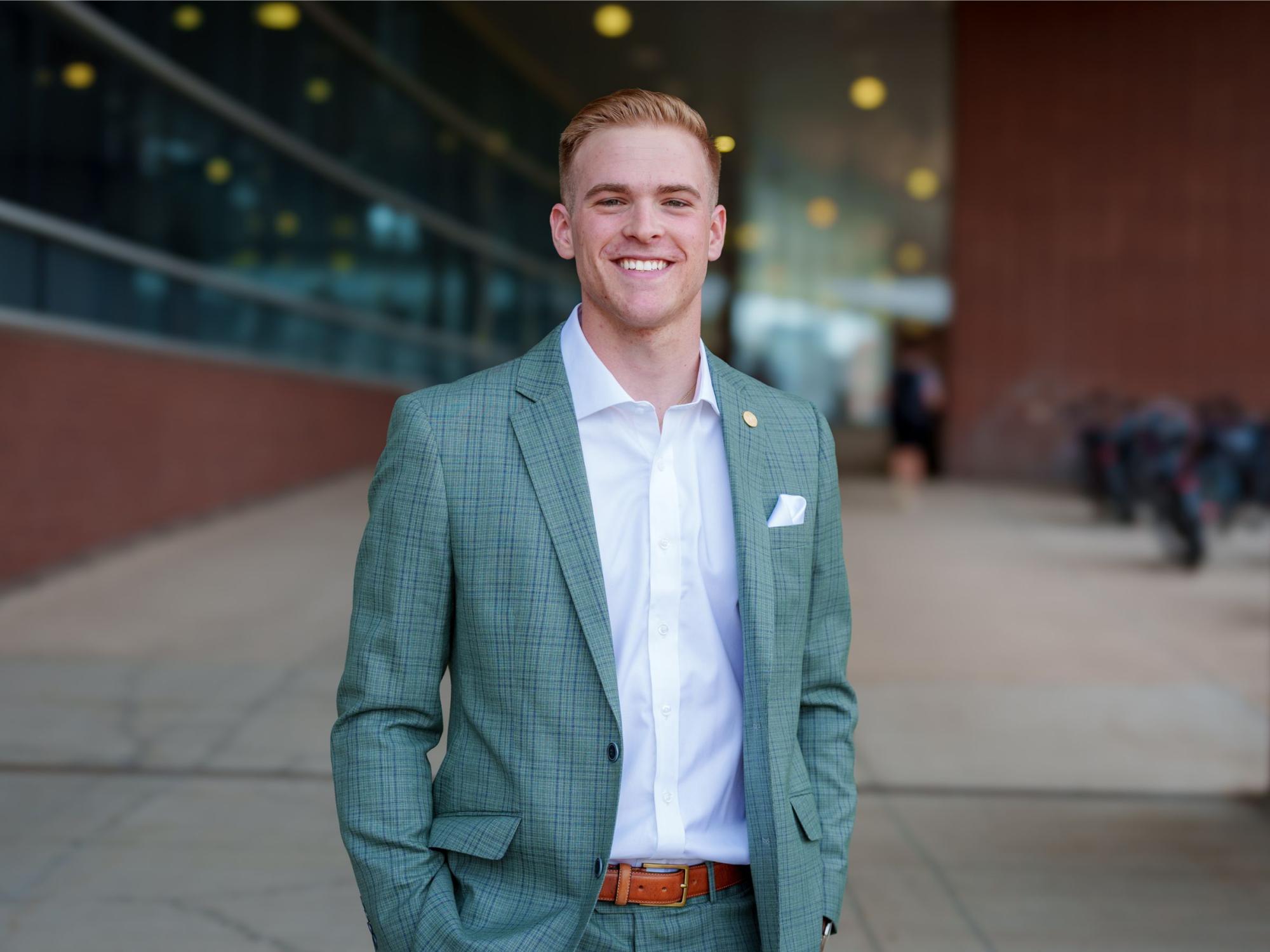 smiling fourth-year college student in suit jacket with campus building in background
