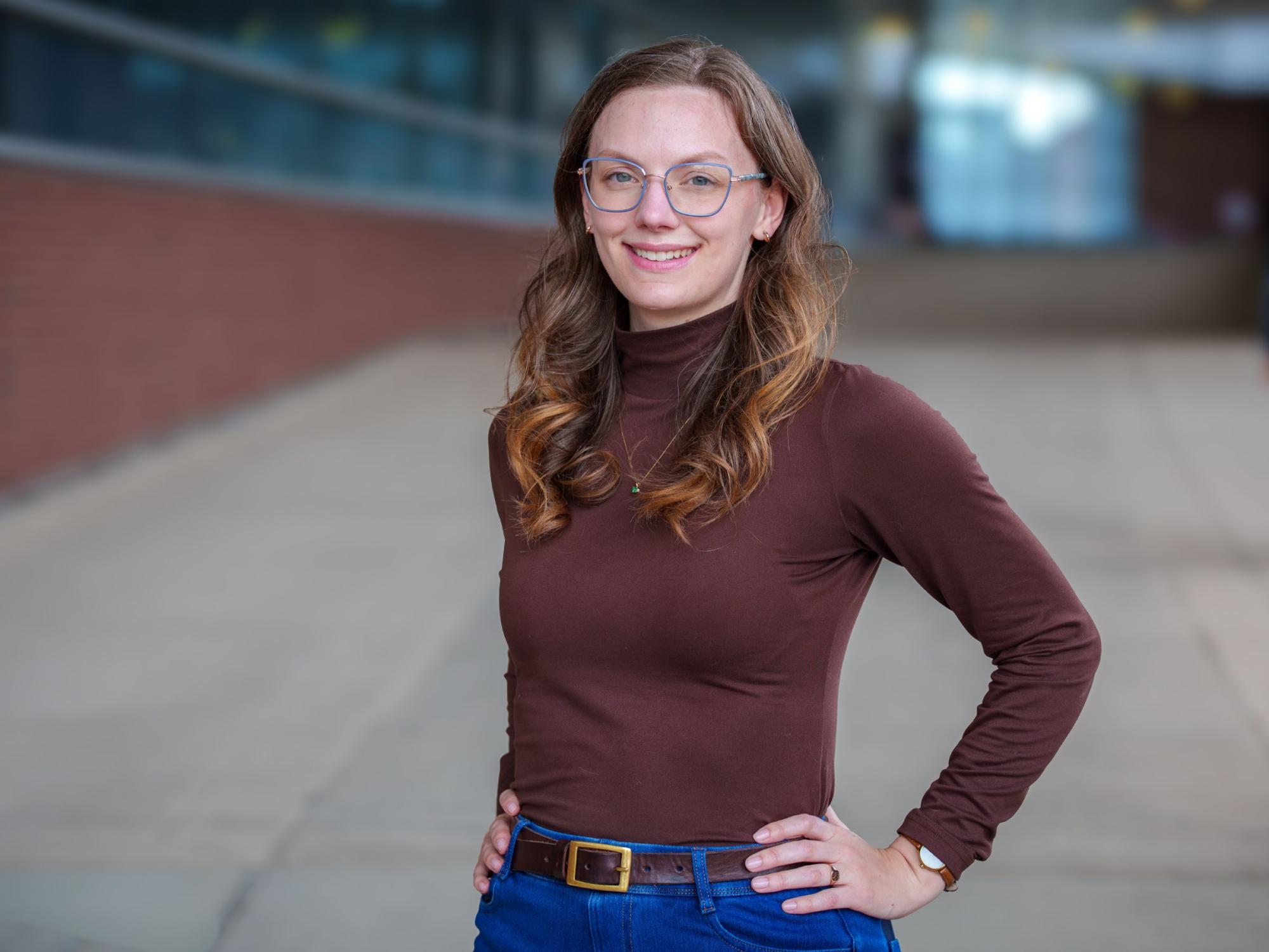person with long hair and glasses stands with hands on hips in front of brick and glass building