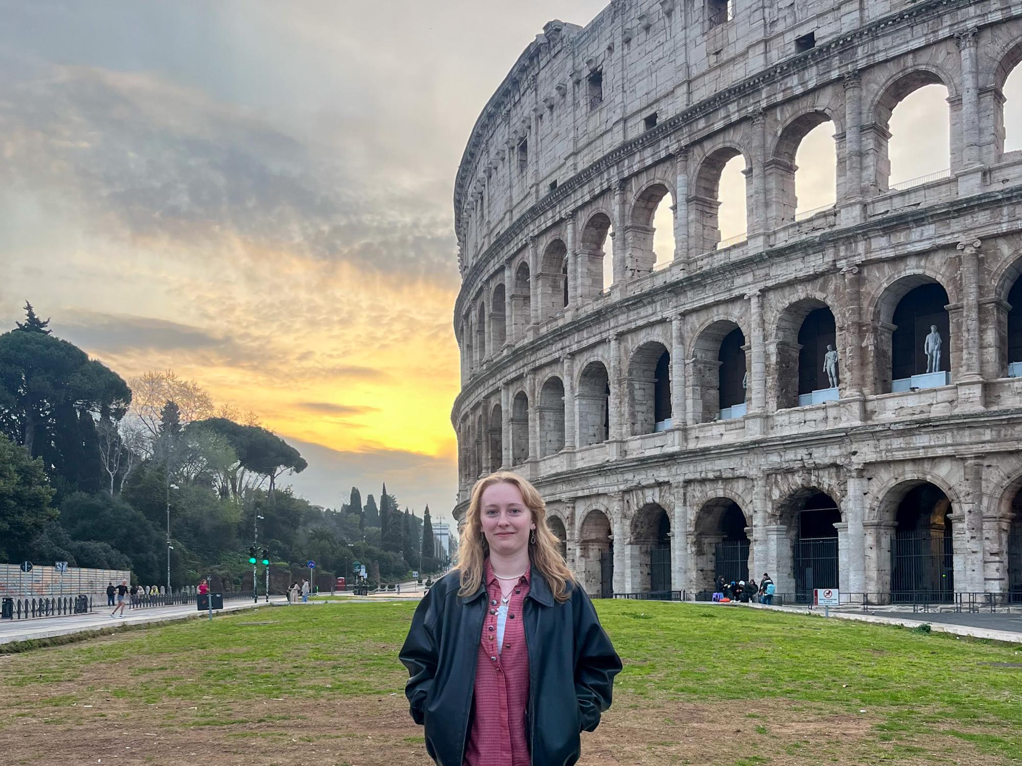 Veronica Swartz wears a red top, black leather jacket, and jeans. She stands in front of the Colosseum in Rome.