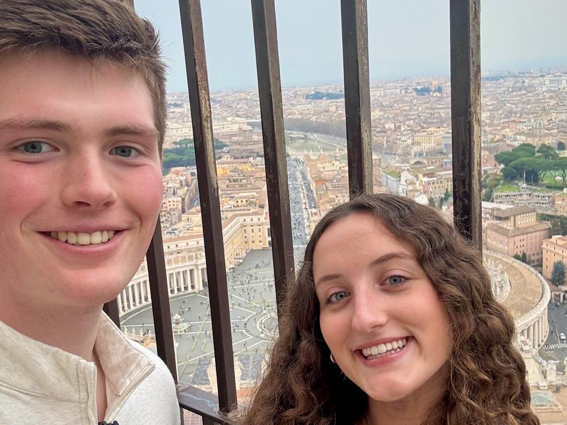 Jacob Ott wears a tan shirt and sunglasses on his shirt. Olivia Mrazik wears a navy shirt and black leather jacket. They stand above the square of the Vatican.