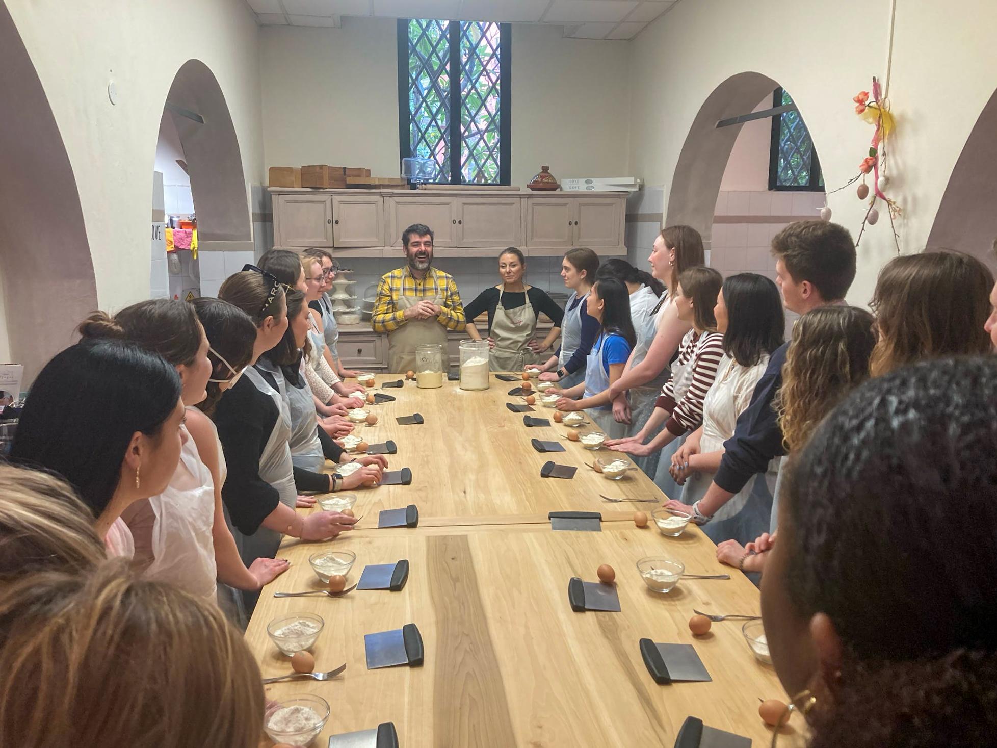 Students stand on either side of the table while their instructor prepares them for the pasta-making class.. 
