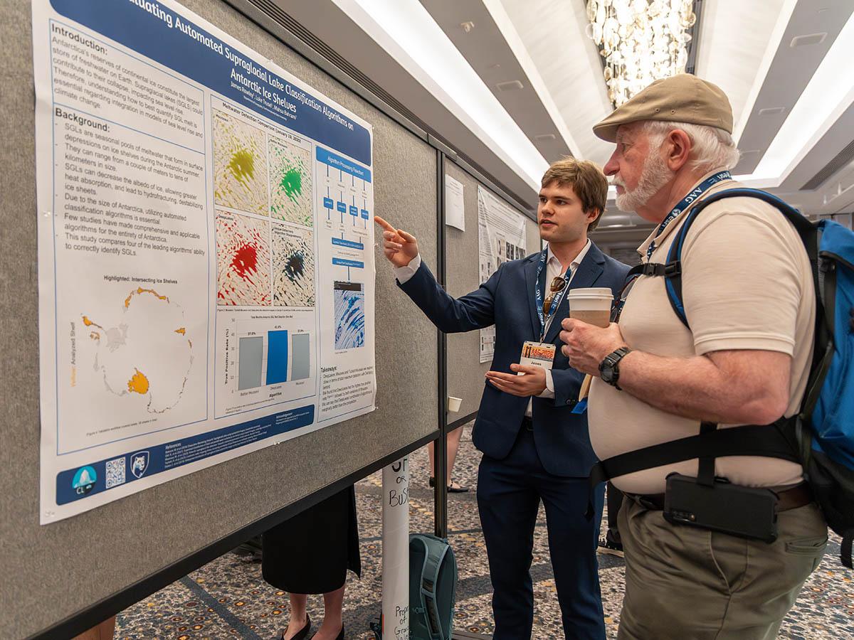 James Rowley in a suit explains a scientific poster to Stuart K. McFeeters, a man with a cap and backpack in a conference setting.