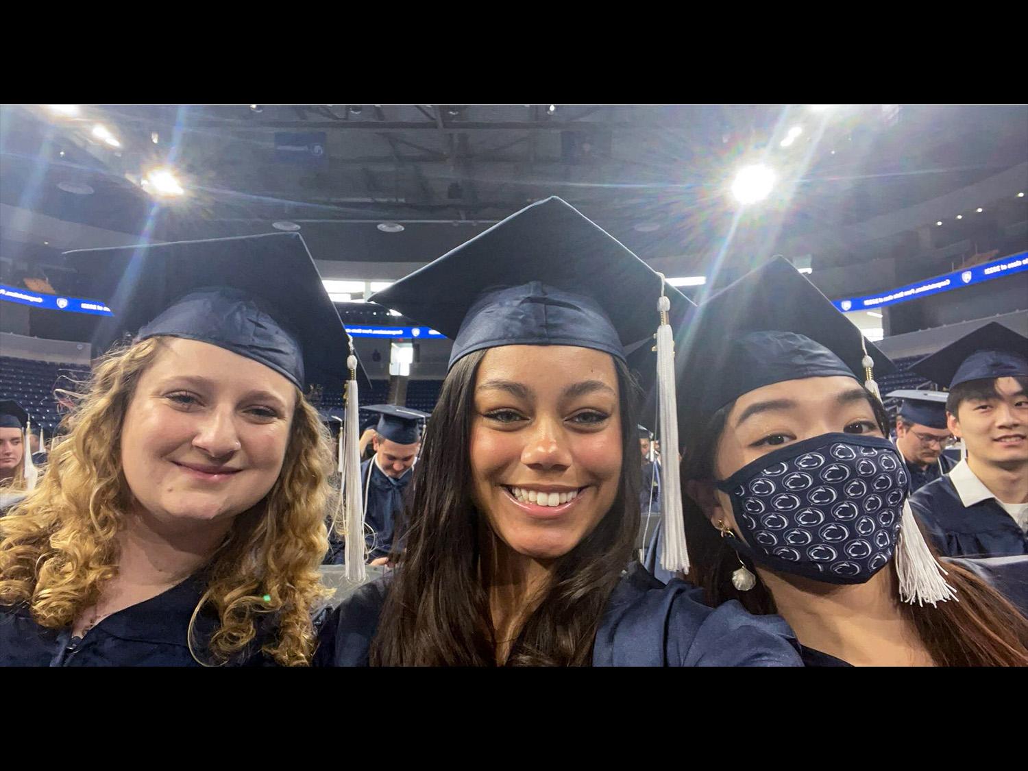 Graduates in caps and gowns smile at a ceremony; one wears a mask. The setting is a brightly lit indoor venue.