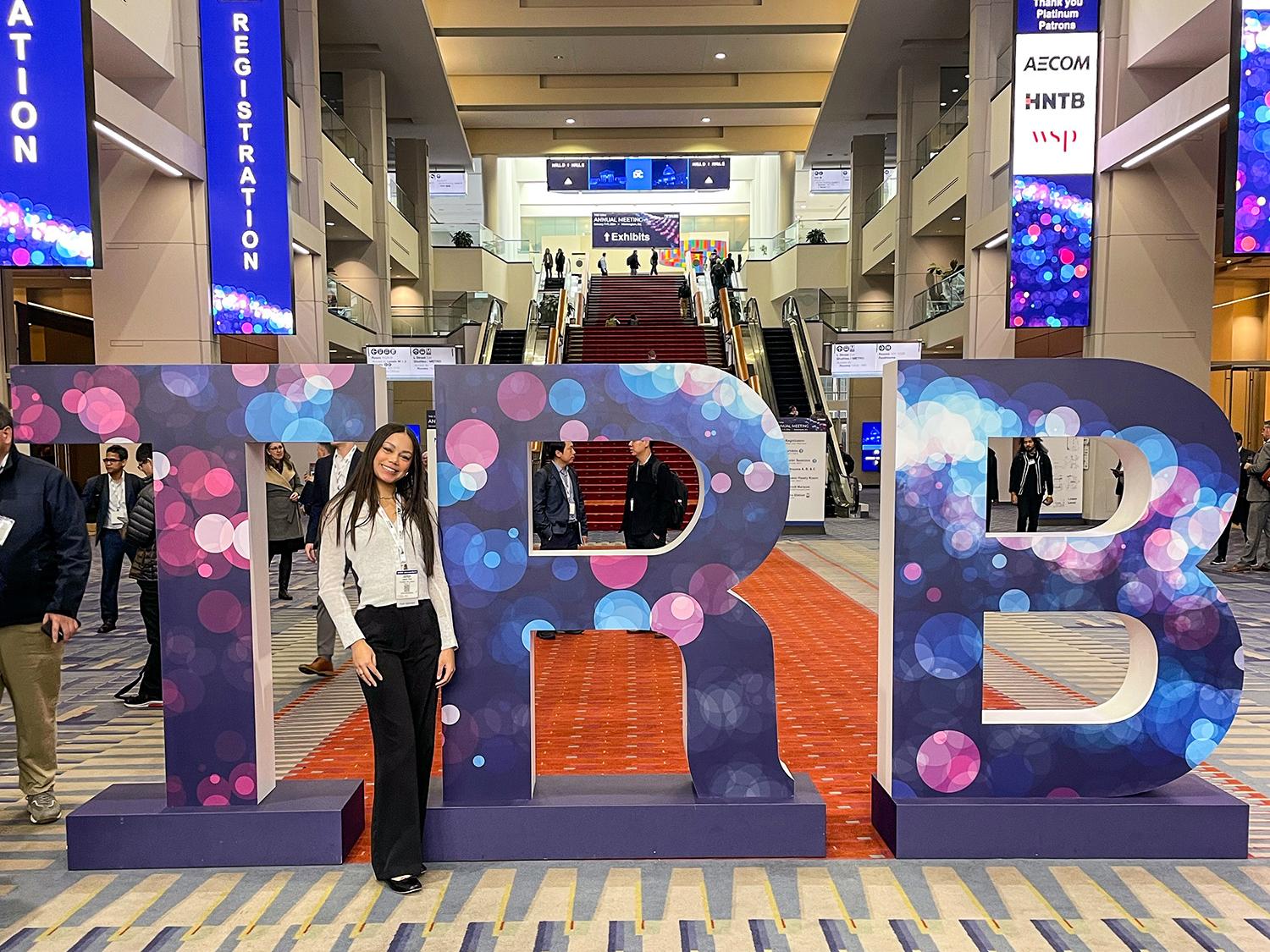 Jazmun stands smiling beside large letters "TRB" at a conference entrance. The scene is lively, with people around and registration signs visible.