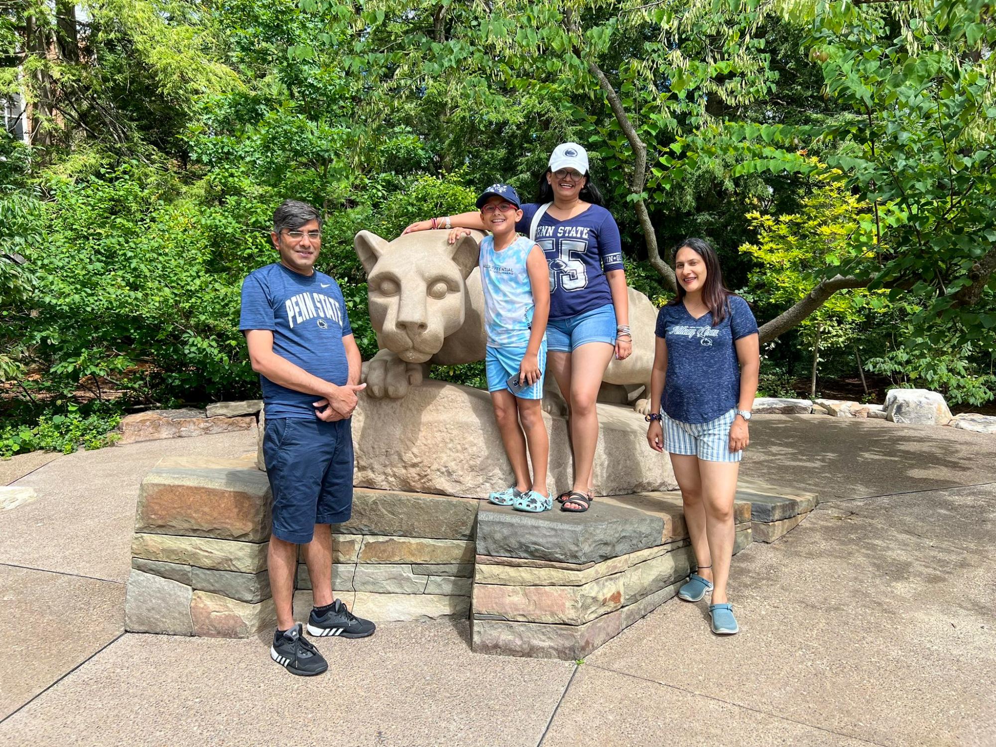 Four people in Penn State shirts pose in front of the Nittany Lion Shrine outdoors among trees.