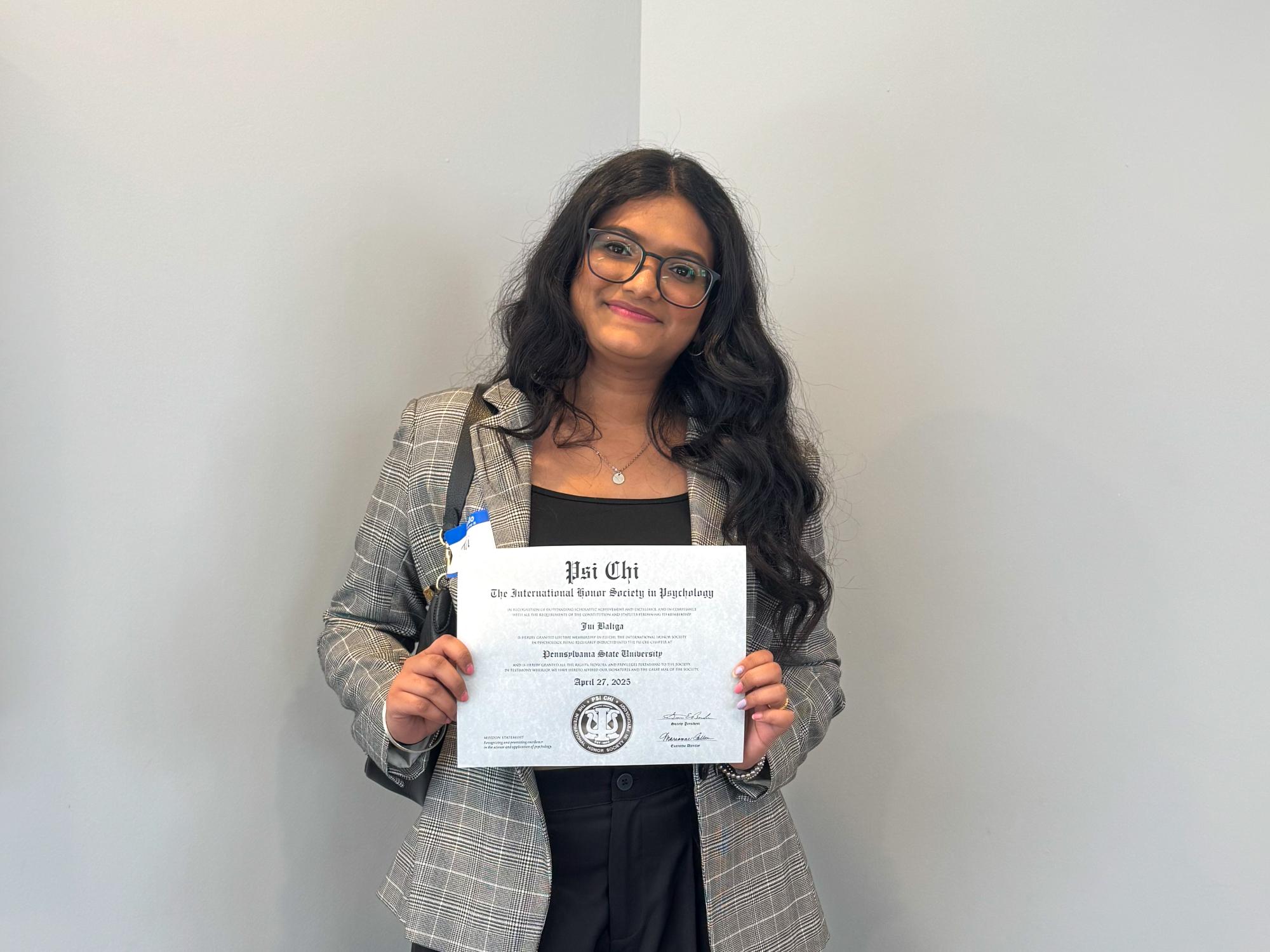 Jui Baliga wears a gray plaid blazer and holds a Psi Chi certificate while standing against a light gray wall.