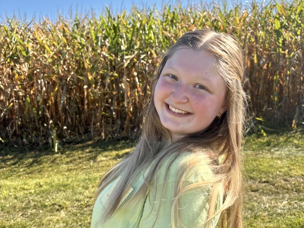 A young woman with long hair poses in front of a corn field.