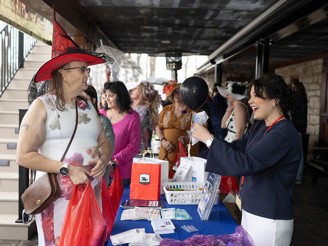 A woman wearing a witch’s hat talks to a smiling woman at a table filled with cards and brochures. Behind them is a group of women.