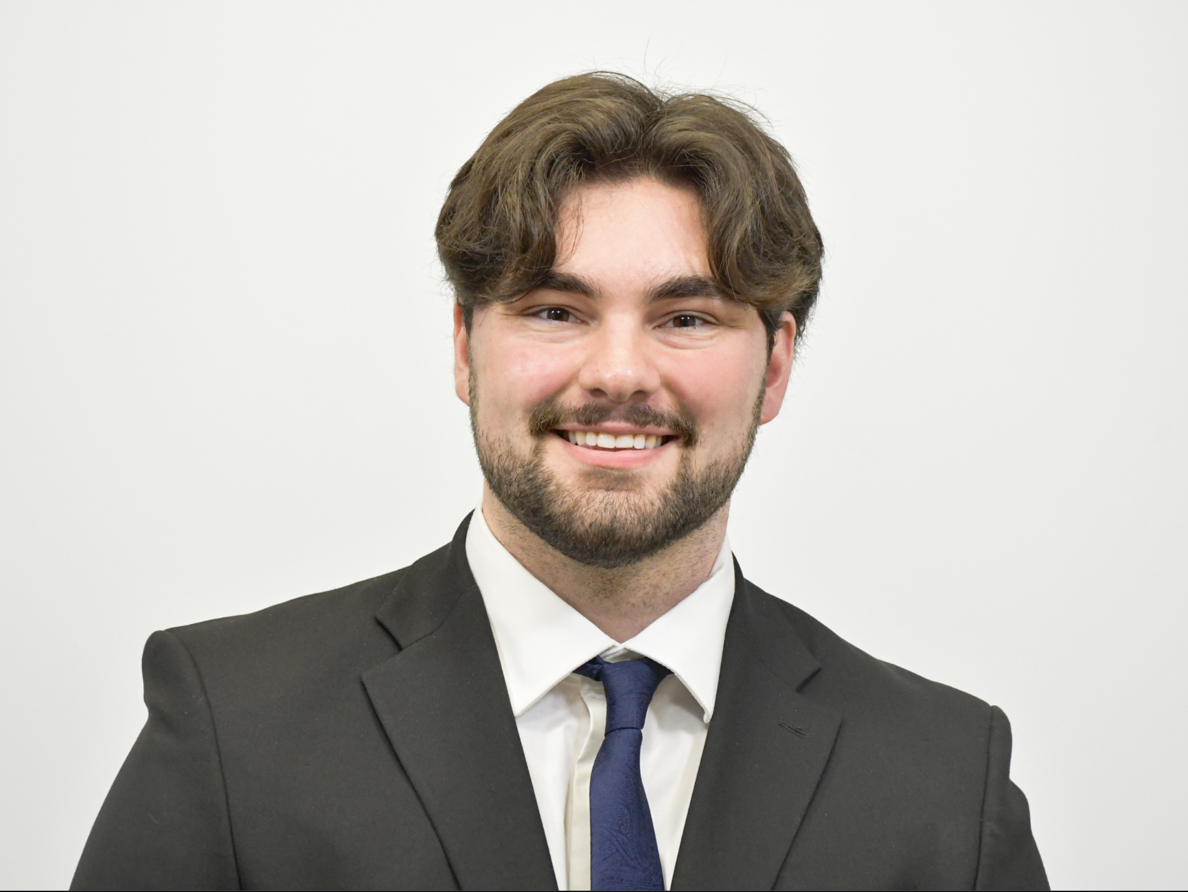 A young man wearing a dark gray suit with white shirt and dark tie.
