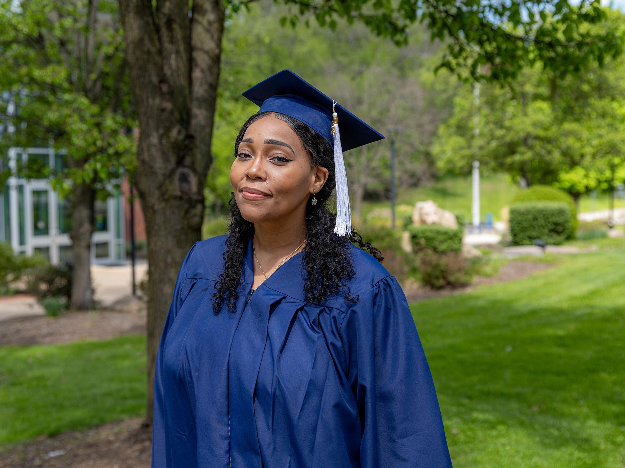 A graduate wearing a blue cap and gown stands outdoors on a campus lawn with trees and a building in the background.