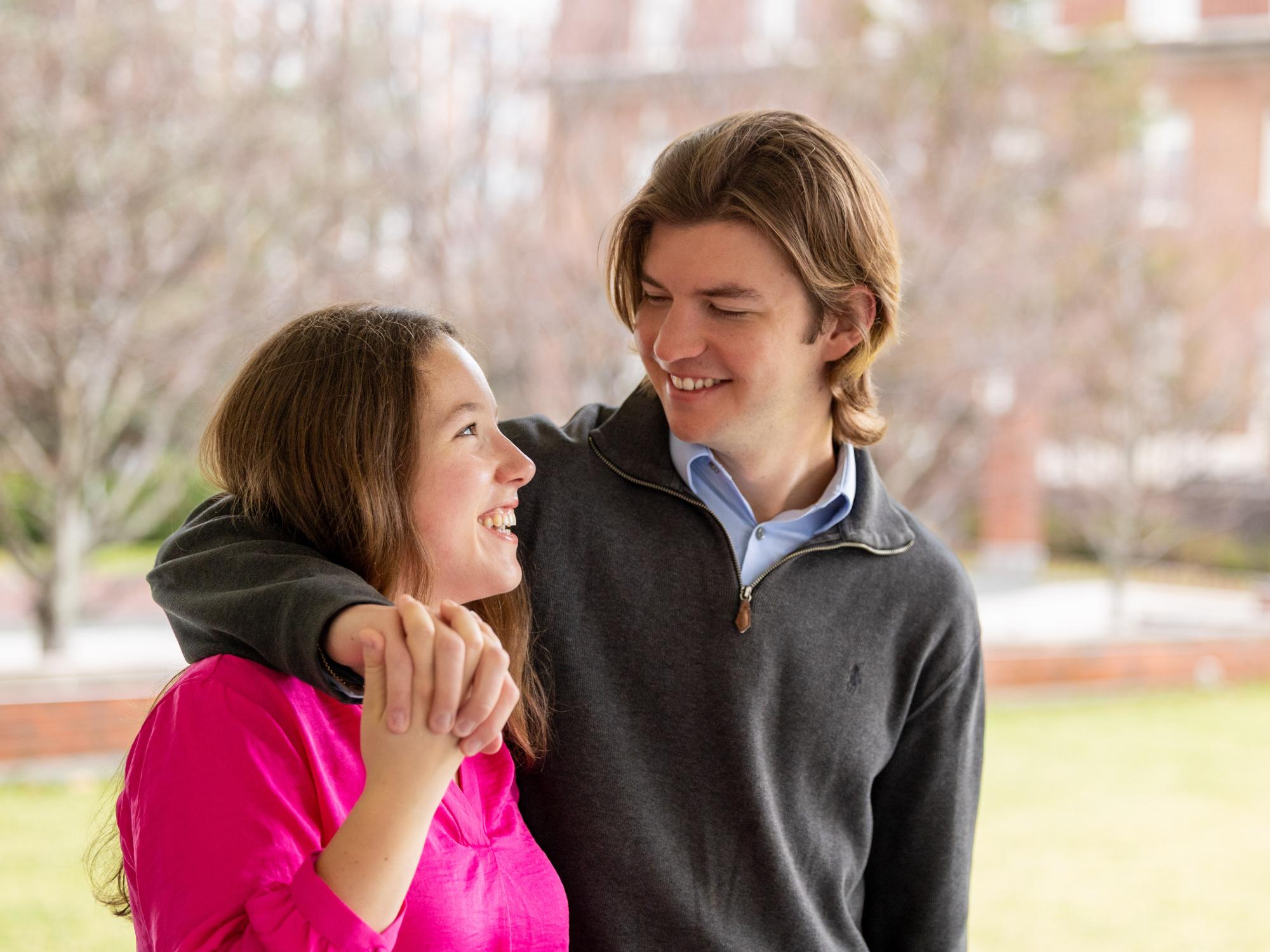 Sabrina, wearing a hot pink blouse, and Morgan, wearing a dark gray quarter-zip and blue button-up, walk together outside holding hands. 