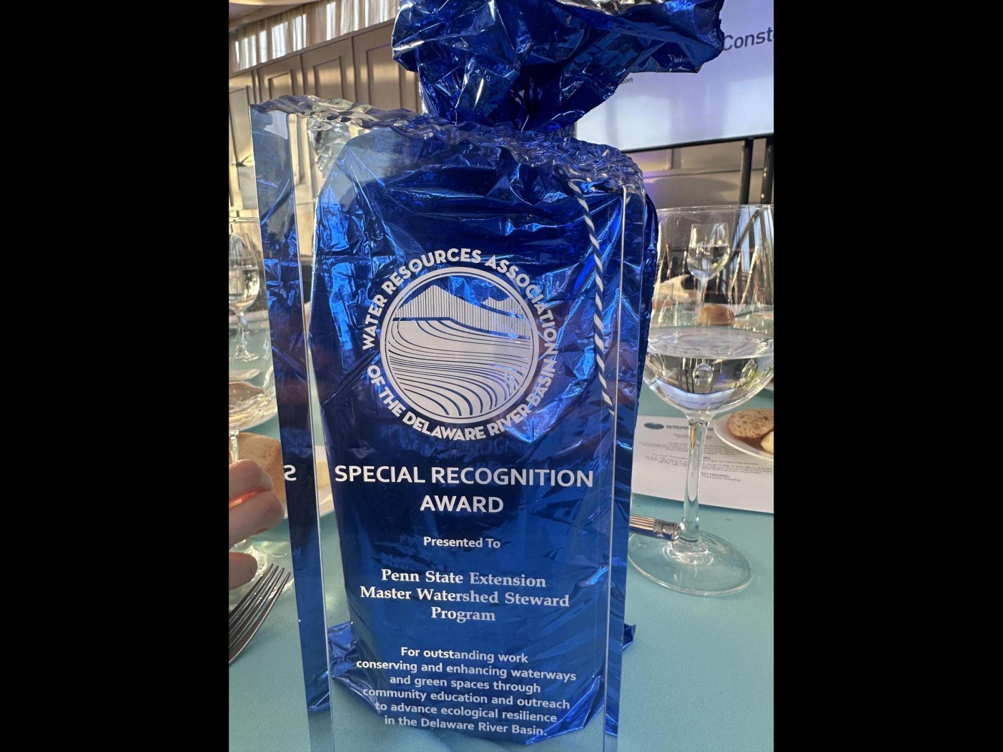 A clear glass award sits on a table set with wine glasses and bread. The award reads “Water Resources Association of the Delaware River Basin – Special Recognition Award,” presented to the Penn State Extension Master Watershed Steward Program for outstanding work conserving and enhancing waterways and green spaces through community education and outreach.