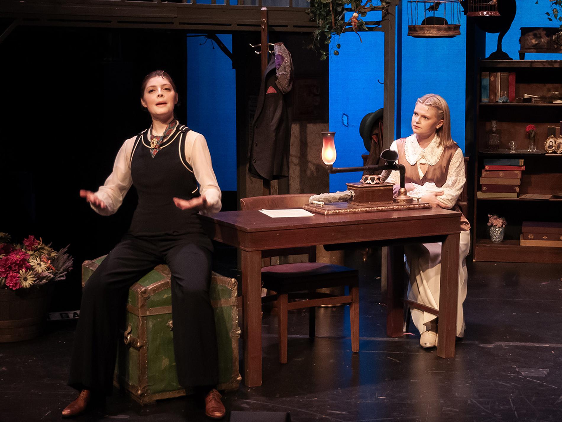 Two actors perform on a stage, seated at a table