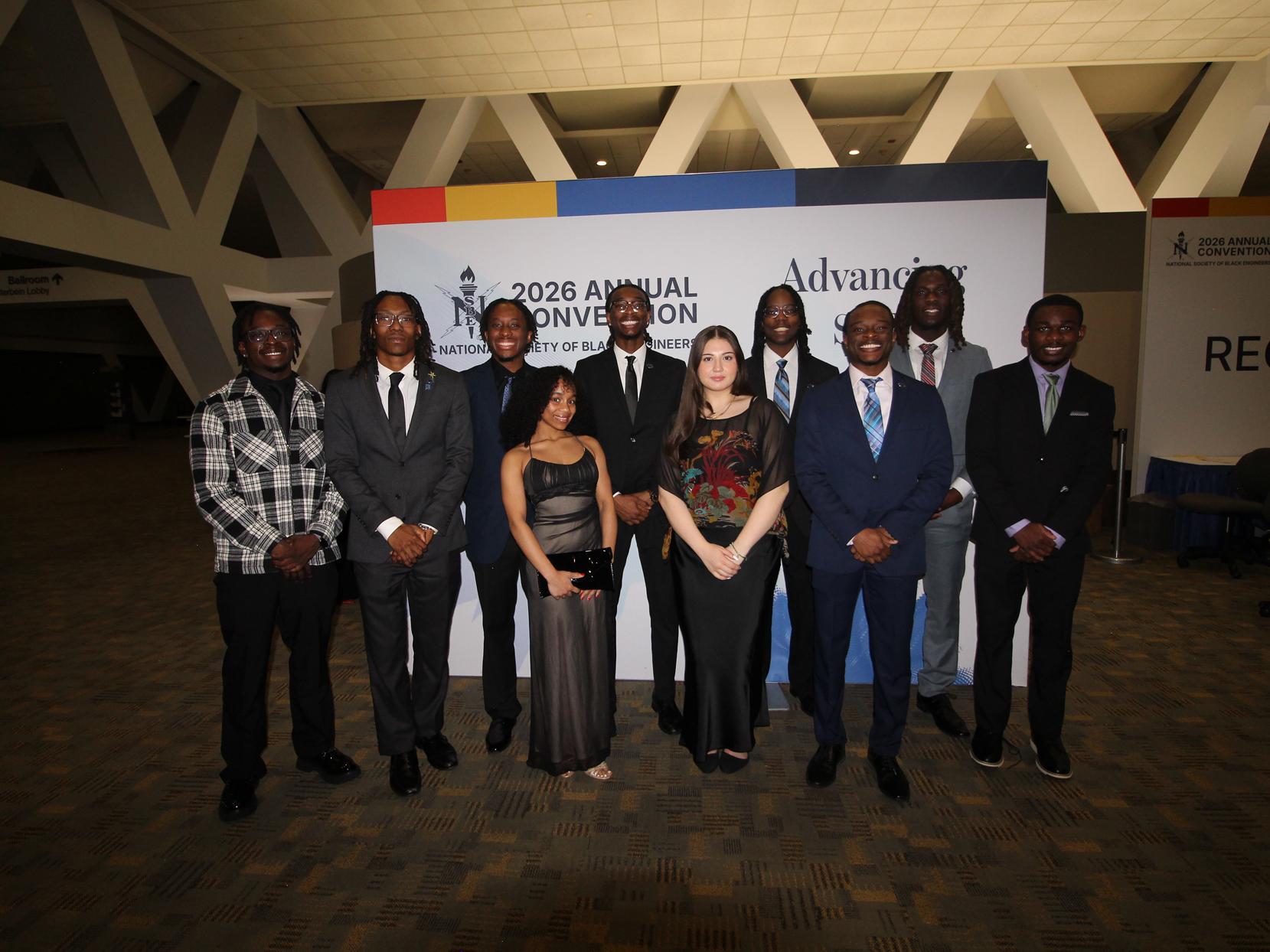 A group of students in formal clothing stand in front of an NSBE convention banner