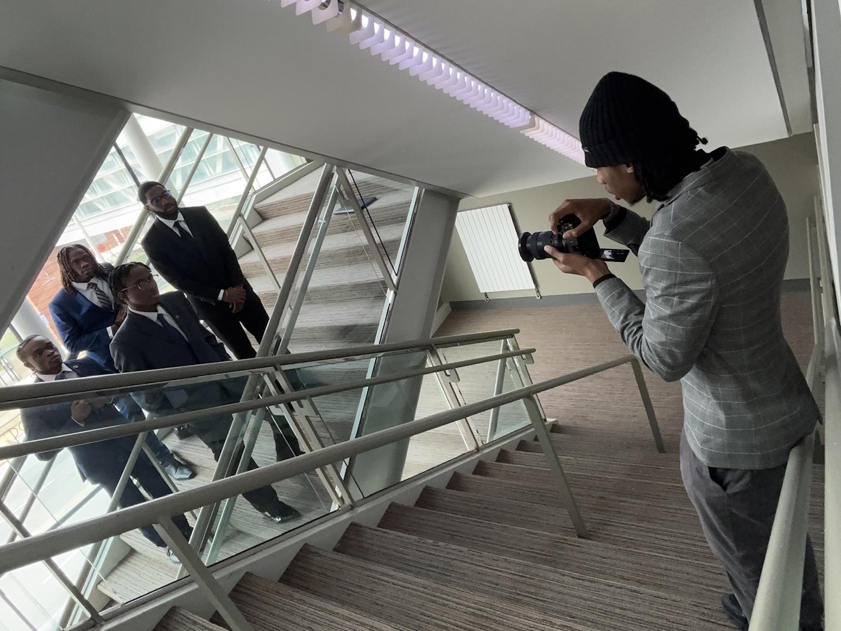 A student holding a camera takes a photo of a group of four students wearing suits in a stairwell