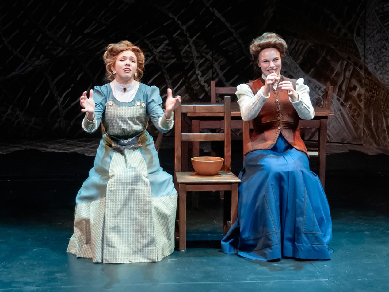 Two women wearing period dresses are sitting next to each other on chairs