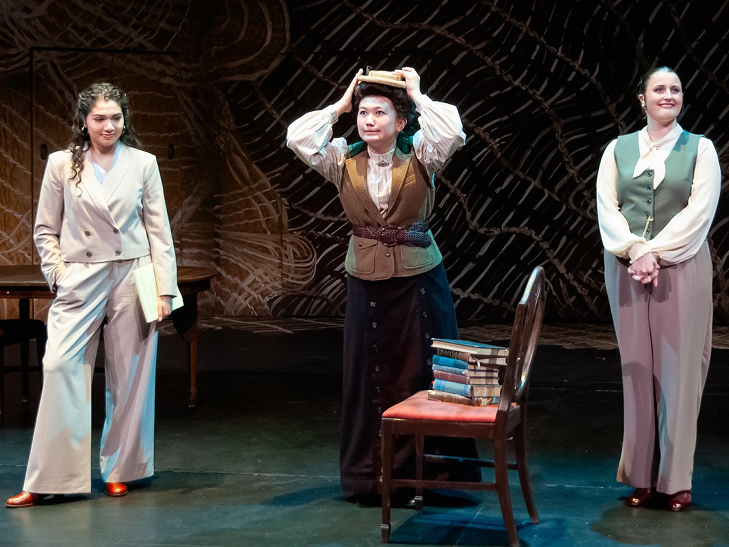 Three actors on a stage in a scene, the middle one is holding a book over her head