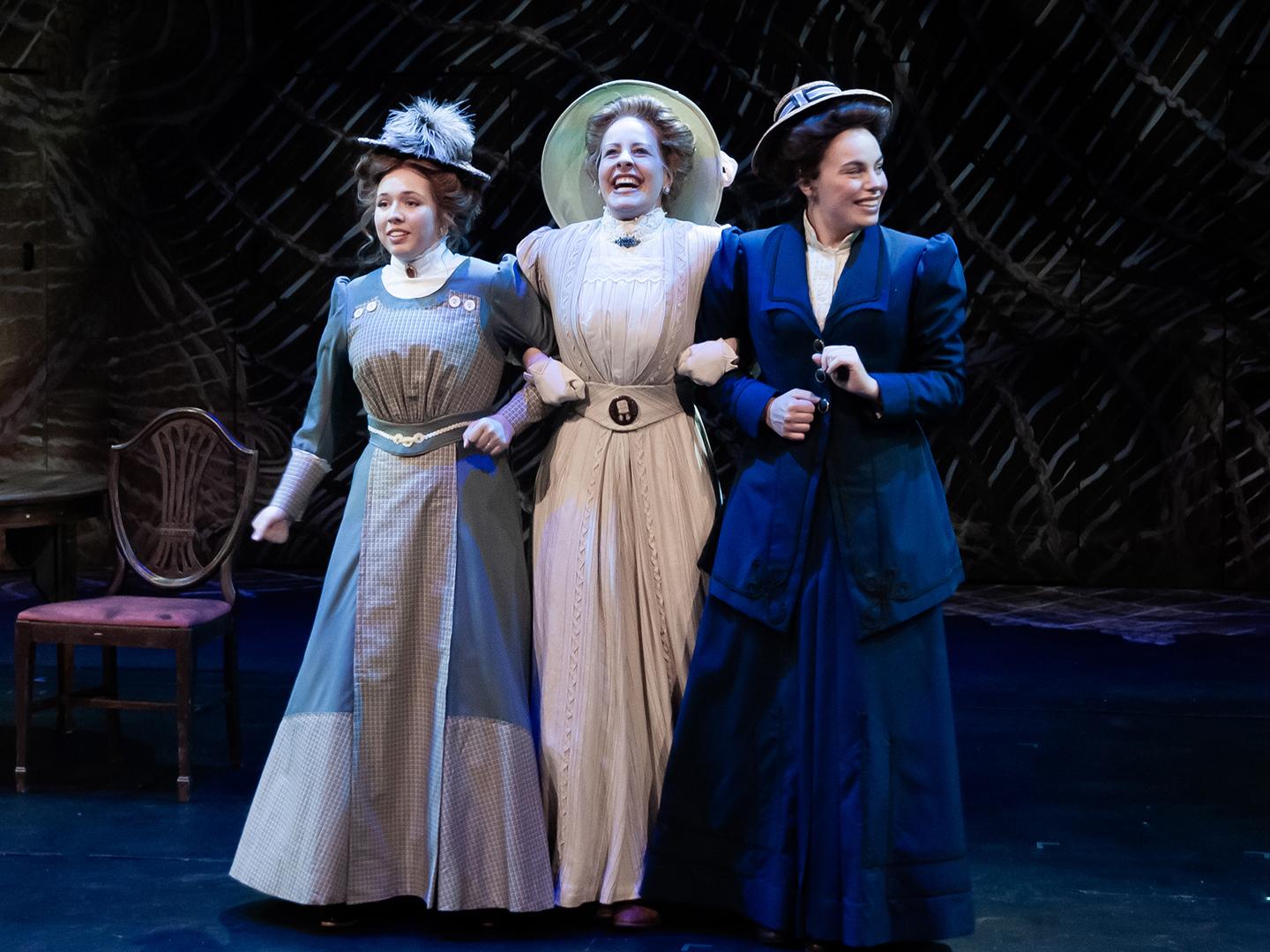 Three ladies in period dresses and big hats walk arm in arm on a stage