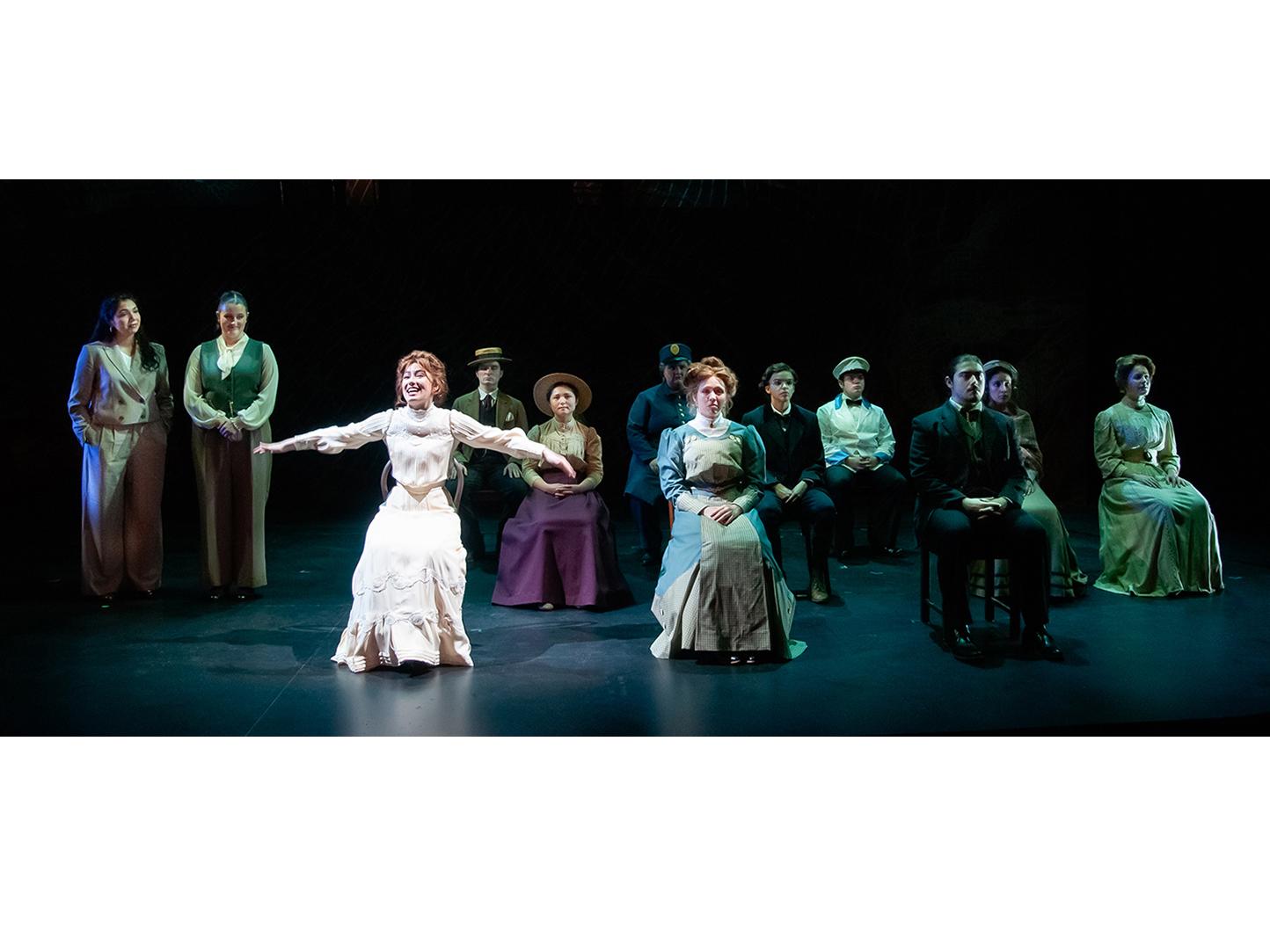 The cast of a theatrical show all on stage together dressed as townsfolk, a smiling woman in the center has her arms spread out wide