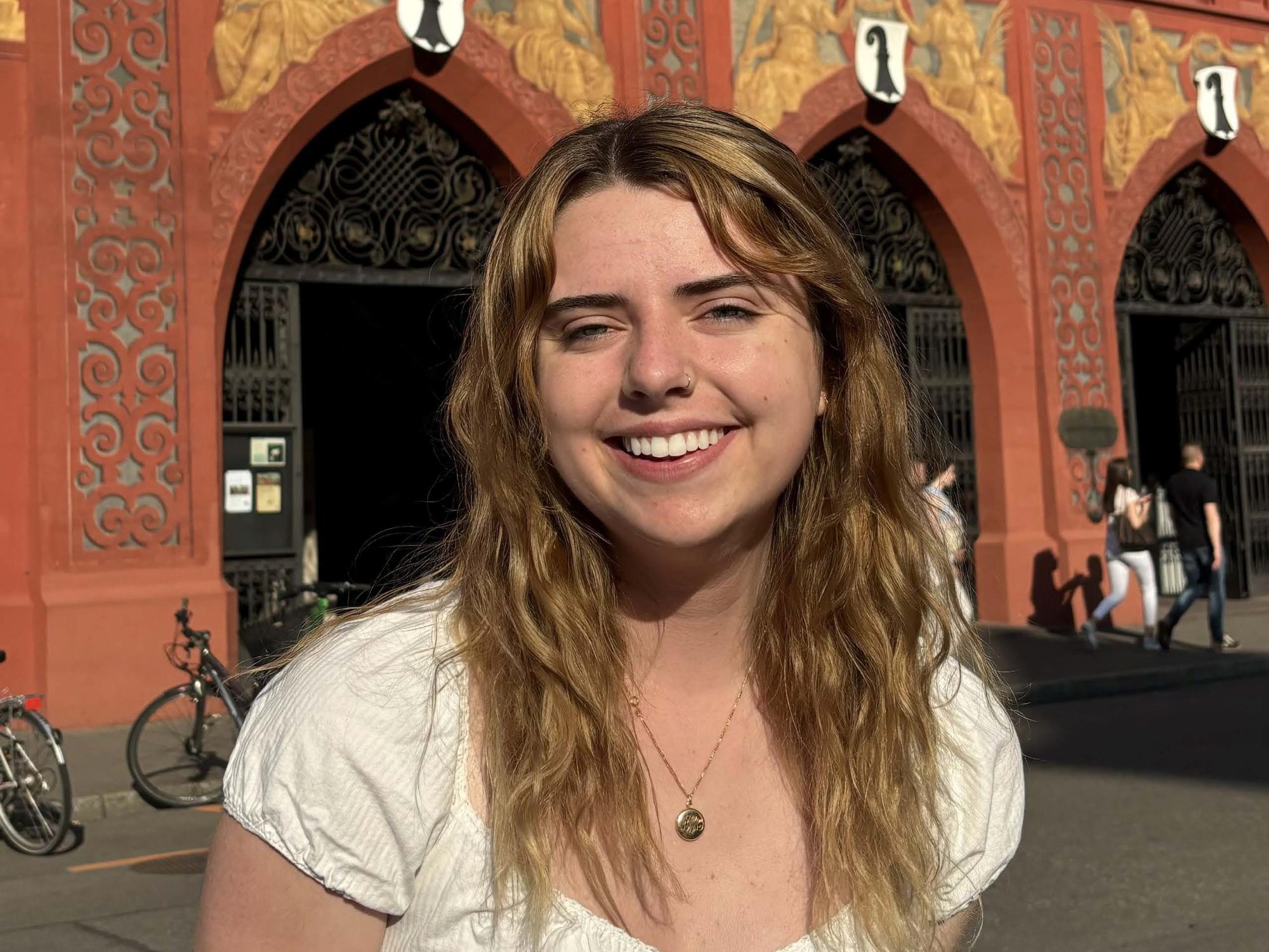 A young woman with long hair and a white shirt stands in front of an arched building.