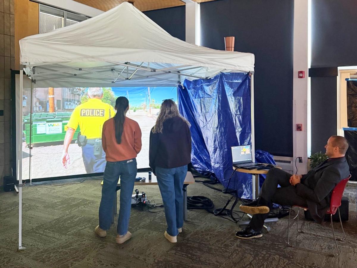 Two participants stand inside a tented simulation setup facing a large screen showing a police scenario, while an instructor sits nearby observing.