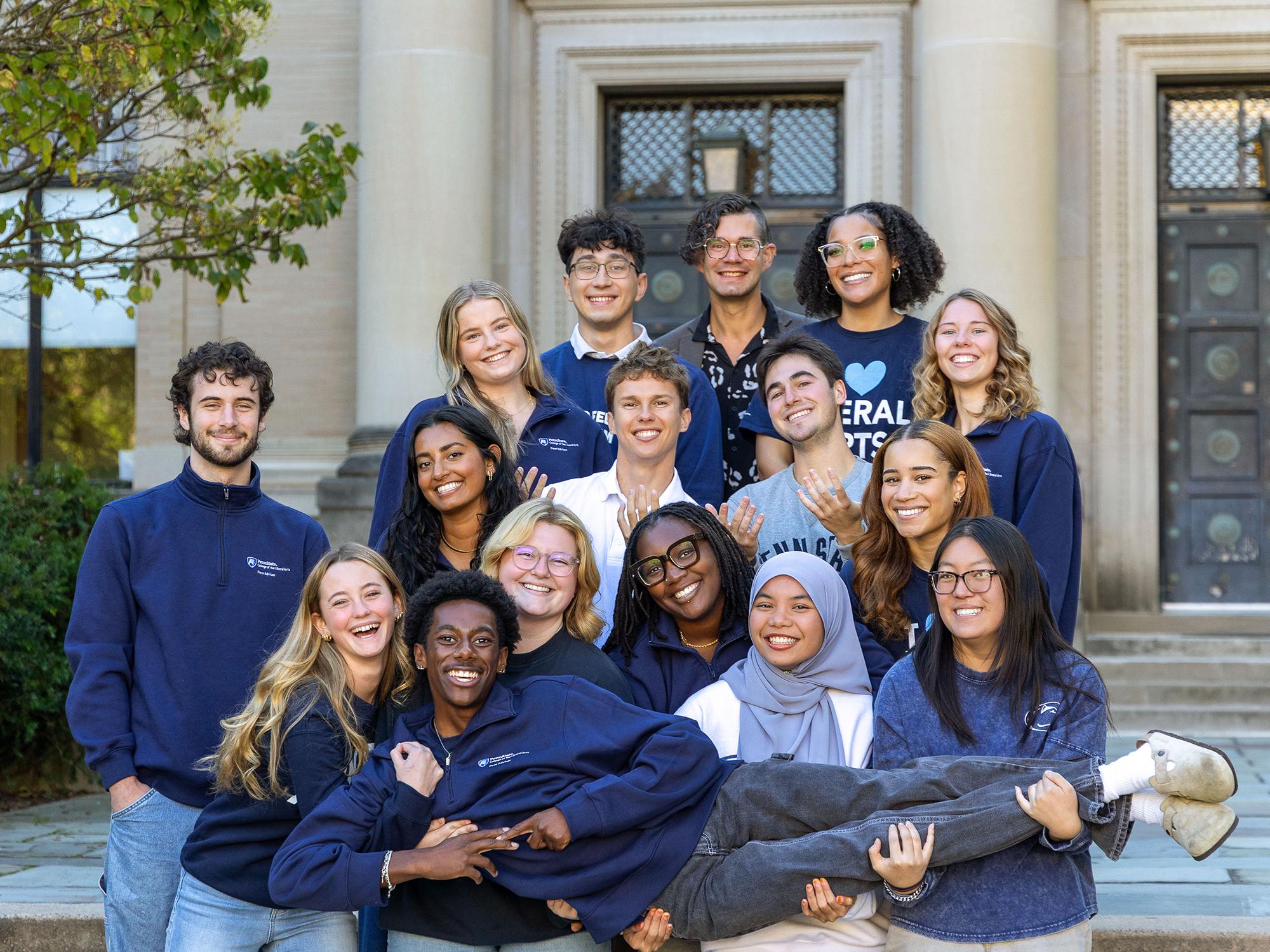 A group of young adults pose together in front of Sparks Building, smiling and leaning in close, holding one person horizontally in a playful pose.