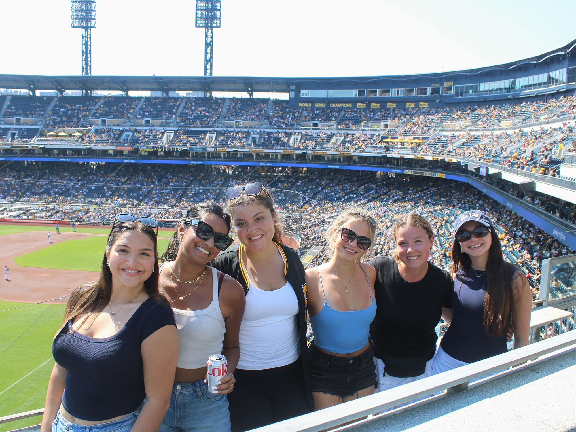 : Six people stand inside a crowded baseball stadium where a game is underway on the field.