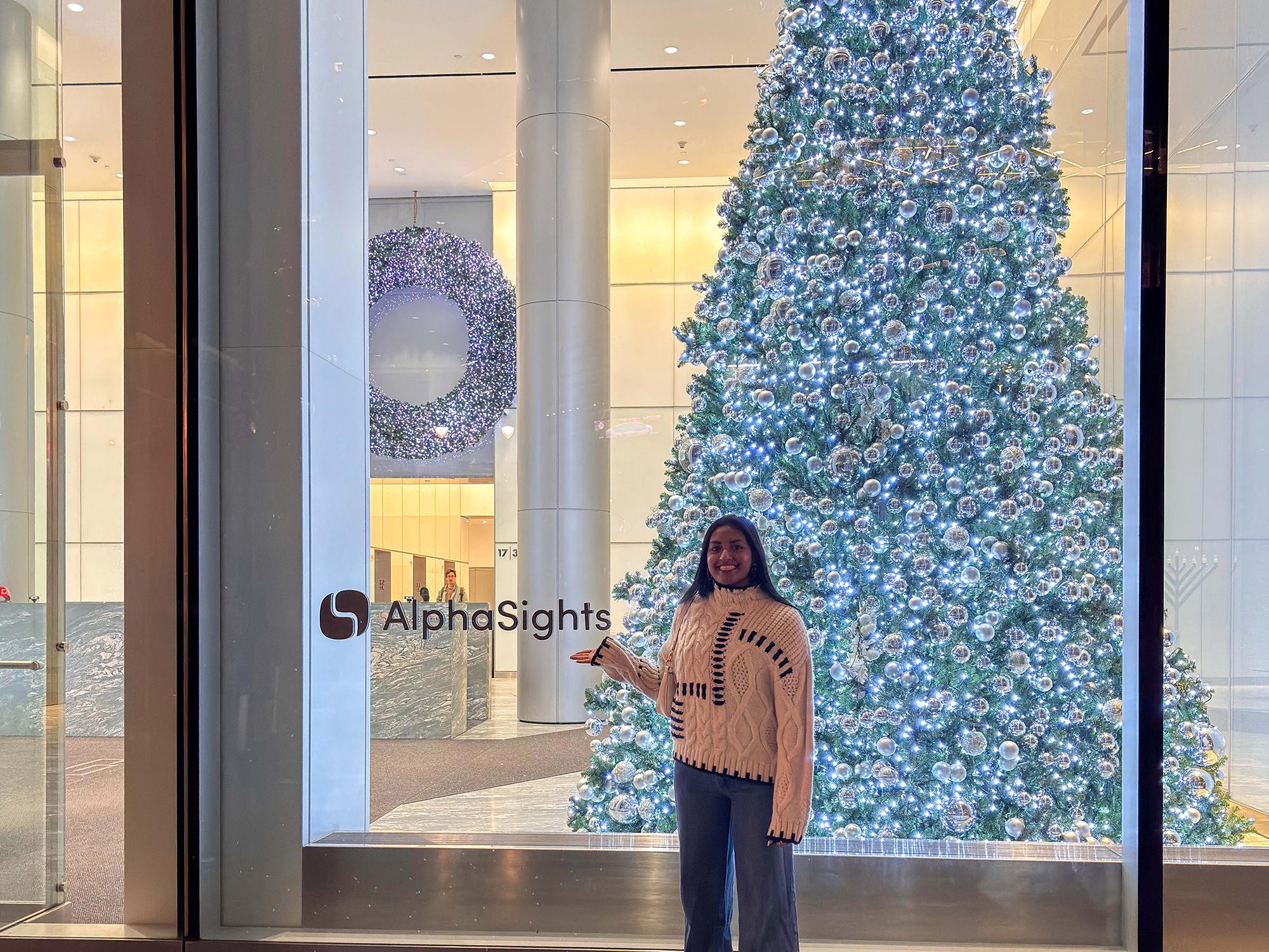 Purani wears a white sweater and stands in front of a large glass window that reads “AlphaSites” which has a decorated Christmas tree lit inside.