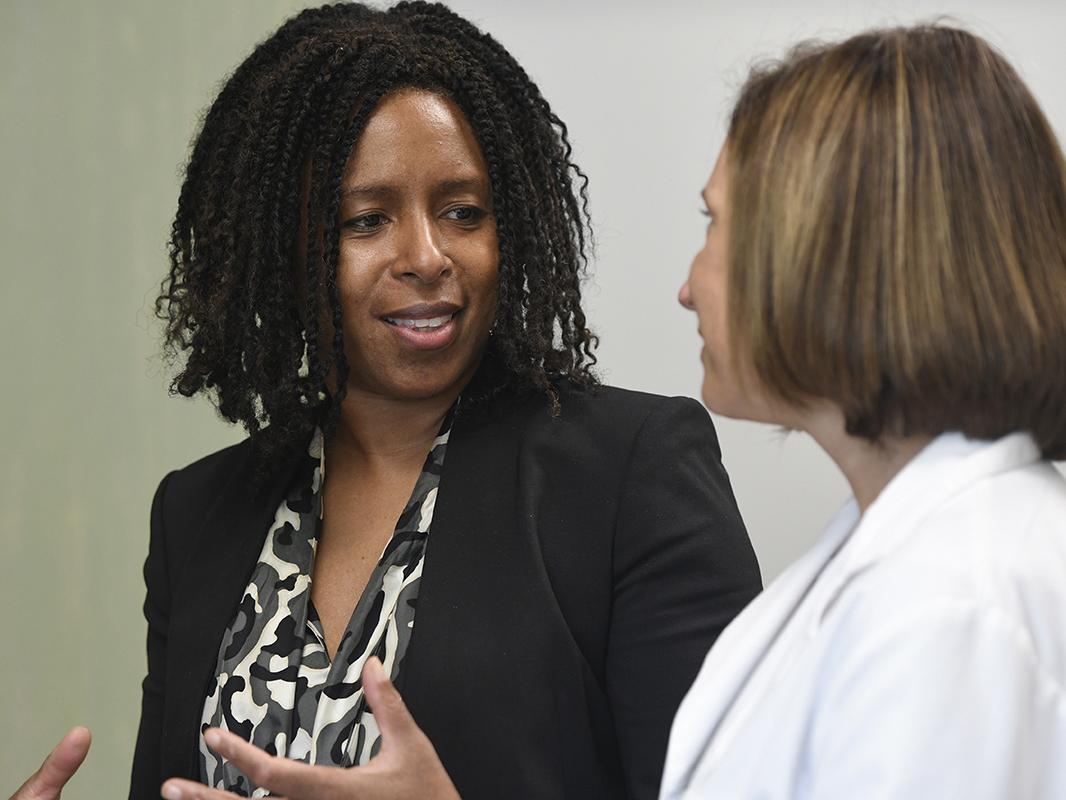 Samantha Butts, left, and Michele Munch of Penn State Health talk in a medical office. Dr. Butts smiles as she gestures with her hands. She has curly hair and is wearing a patterned blouse and jacket. Munch has short hair and is wearing a lab coat.