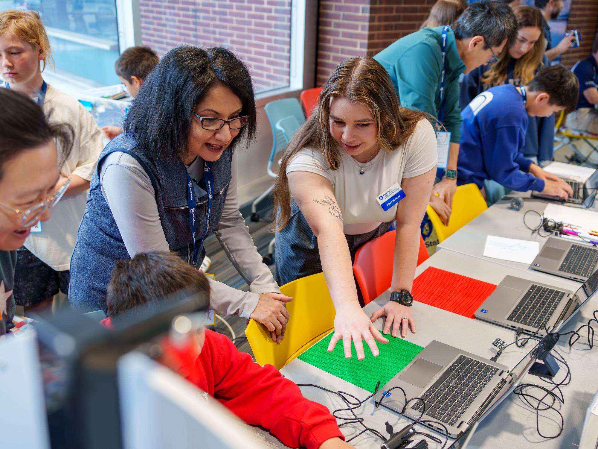 A group of adults and children gather around a table during a hands-on activity. Two women lean over to guide a child seated at the table, pointing and engaging with materials in front of them, including colorful baseplates, laptops, and electronic components.
