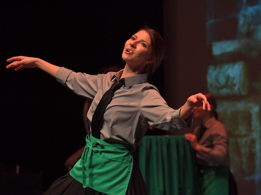 Layna Henry performs on stage wearing a gray shirt, black tie, and green apron with arms extended under stage lighting.