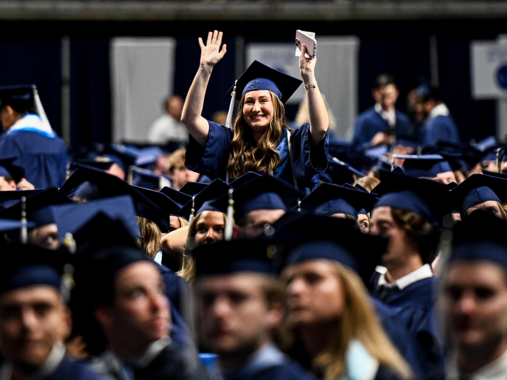 A Penn State graduate in a blue cap and gown stands among fellow graduates and waves to family and friends.