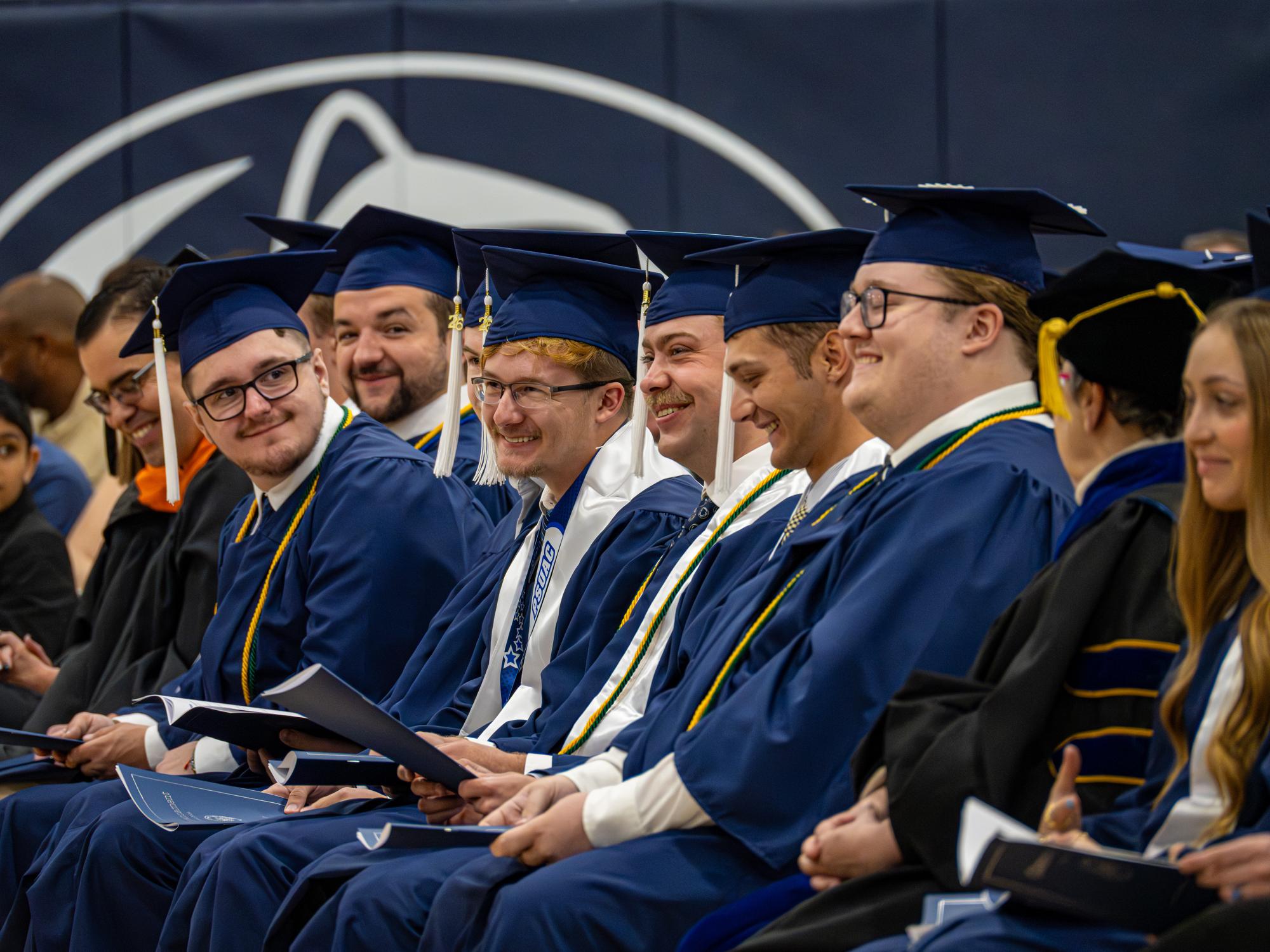 Graduates in blue caps and gowns during Penn State New Kensington’s commencement ceremony, holding printed programs as faculty members in academic regalia join them. Honor cords and stoles are visible among the graduates, with a large Penn State Athletics logo displayed on the wall to the side of the graduates and audience members.