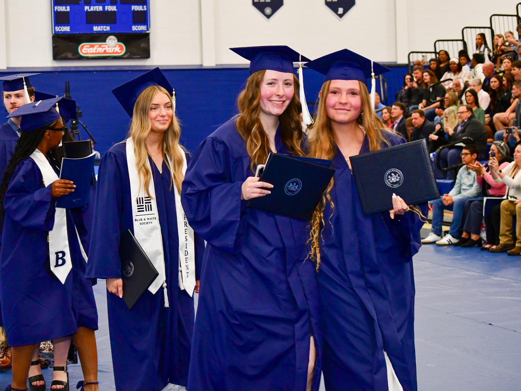 Two students stand together in caps and gowns showing their diplomas while students walk behind them