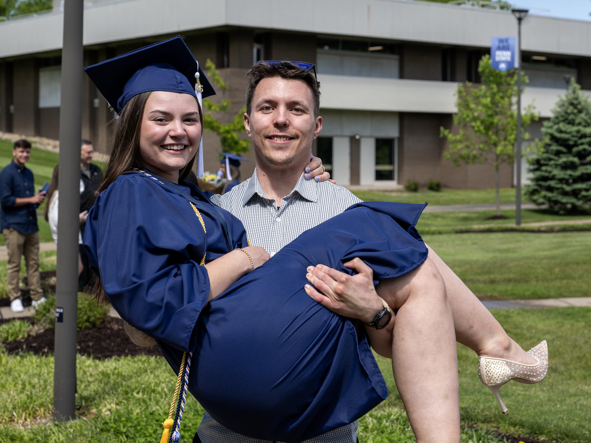 A Penn State Fayette graduate in a blue cap and gown smiles while being held by a man outdoors following commencement. The campus lawn and buildings are visible in the background.