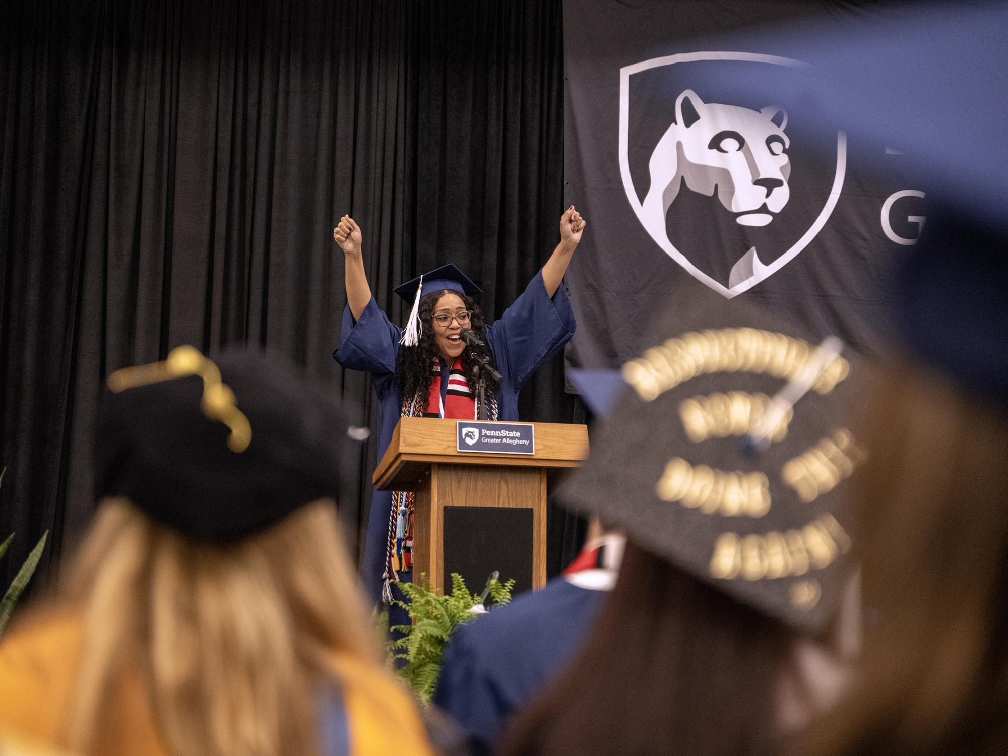 A student commencement speaker stands at a podium on stage wearing a blue cap and gown, raising both hands above the podium. Graduates in caps and gowns are visible in the foreground, and a Penn State Greater Allegheny logo hangs on a dark backdrop behind the stage.