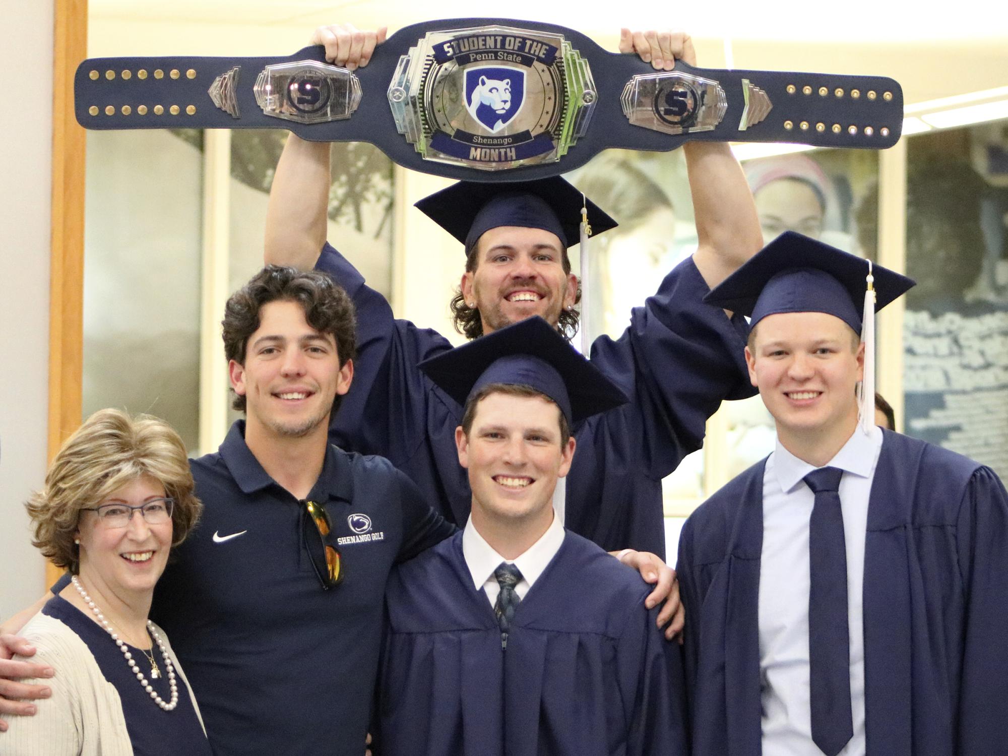 Three graduates wearing blue cpas and gowns pose with two family members as one graduate holds up a large championship belt with the Penn State logo.