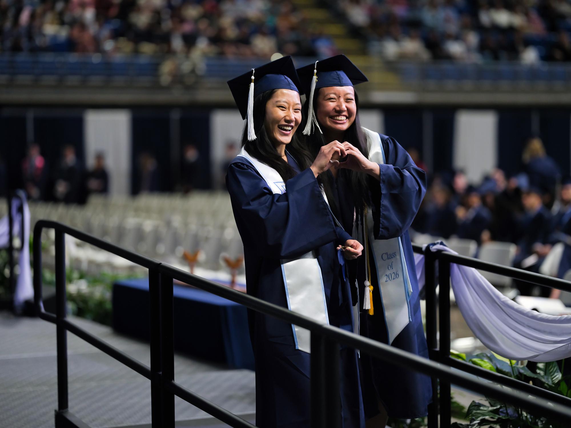 Two students wearing blue caps and gowns make the shape of a heart with their hands