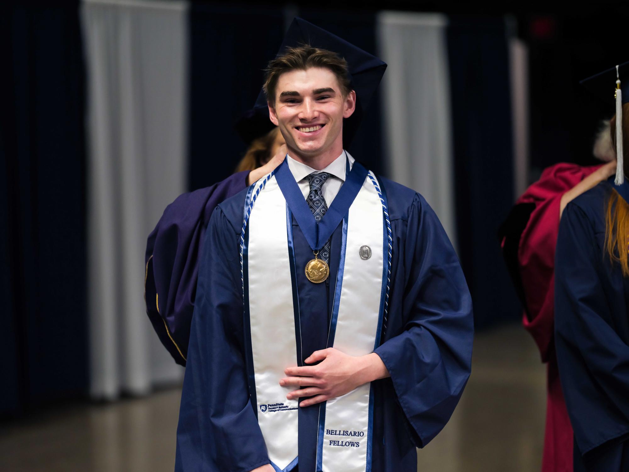 A graduate in a blue cap and gown smiles as someone stands behind them putting a gold medallion around their neck. The graduate wears a white stole with “Bellisario Fellows” embroidered near the bottom and honor cords.