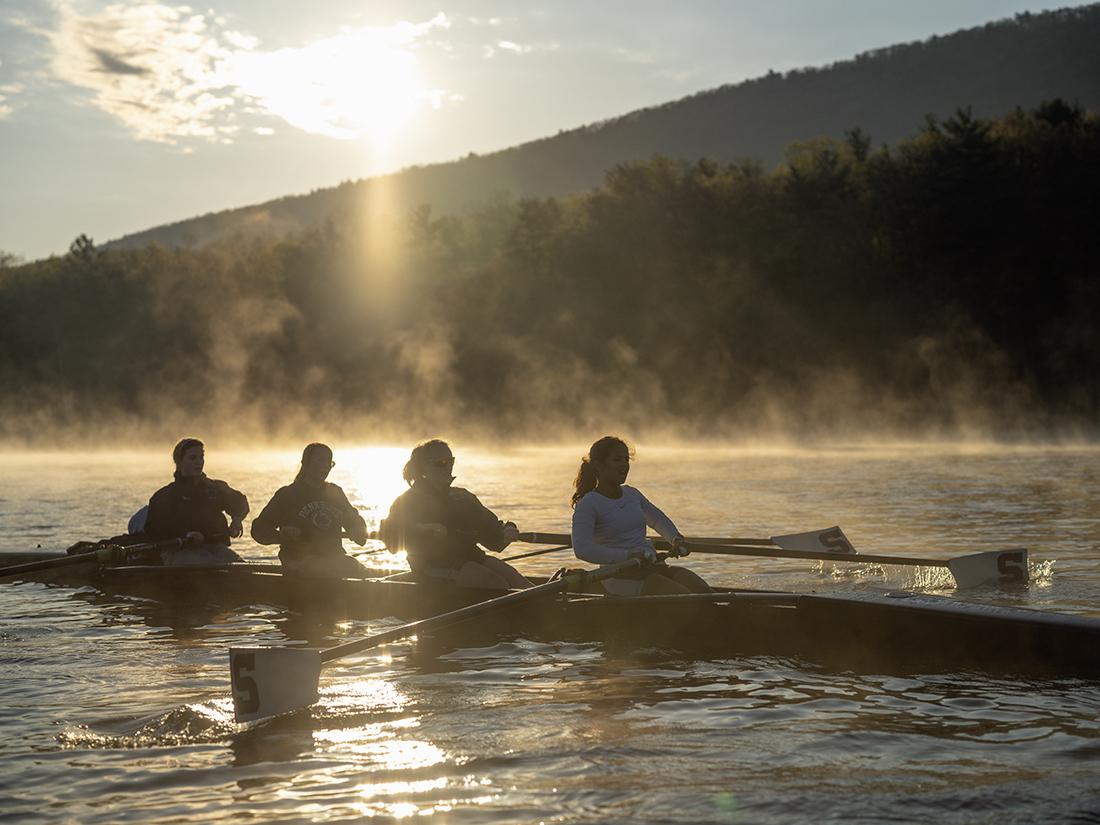 Four members of a crew team in the shadows paddle a shell on a lake at sunrise