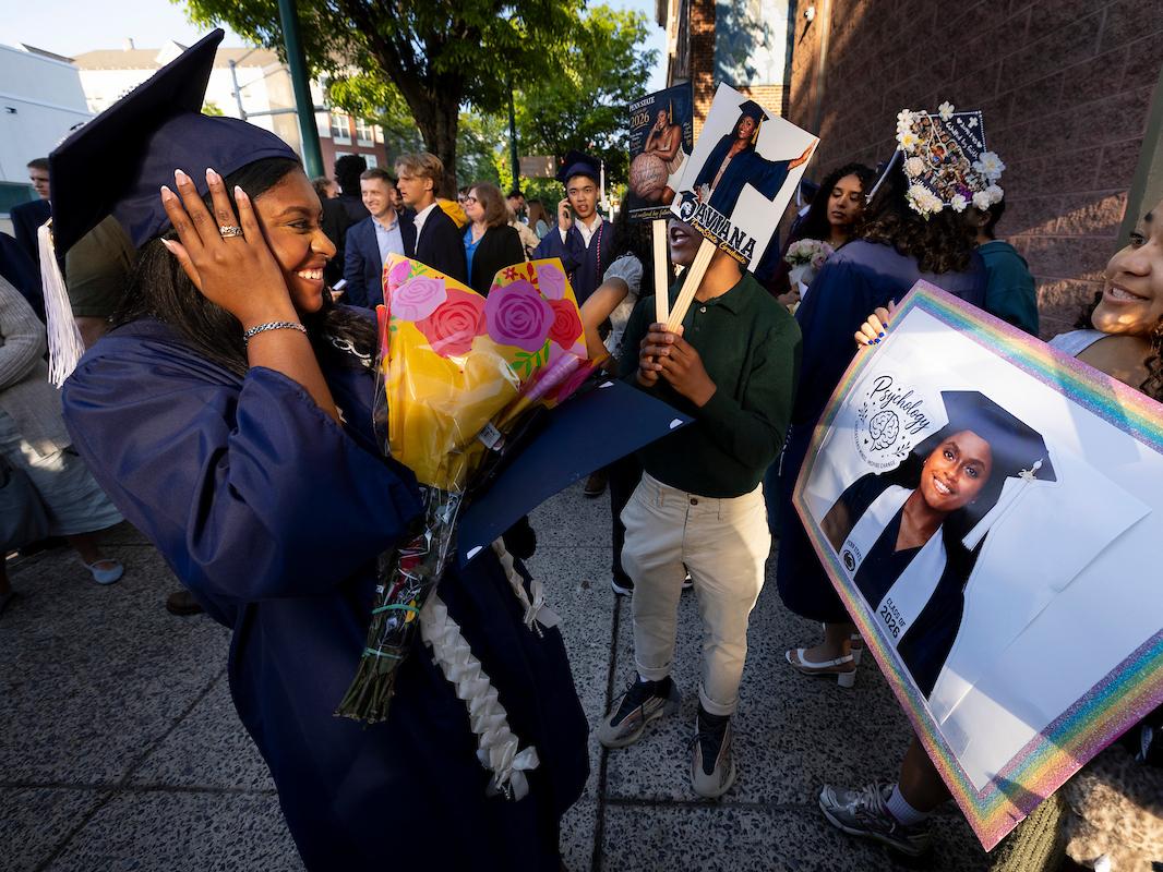 Student in grad cap holding flowers smiles at a family member holding a large photo of her