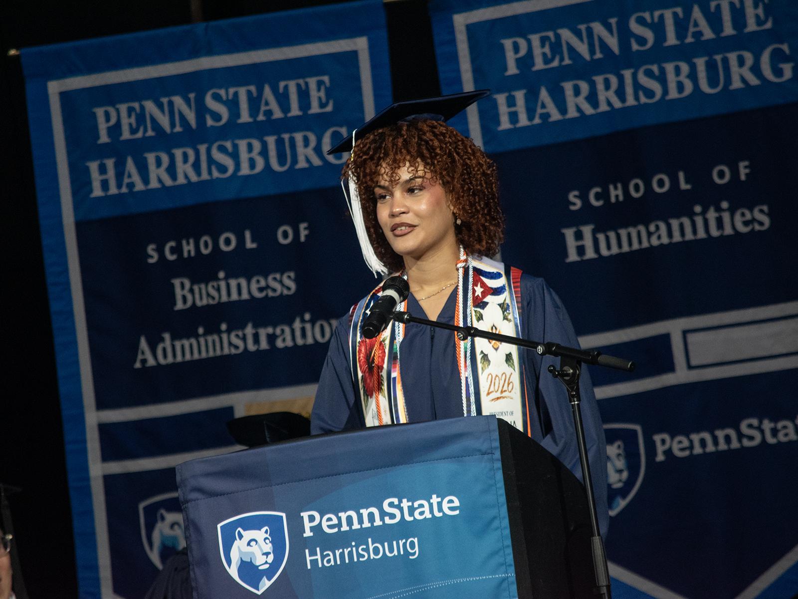 Karyme Font, wearing a cap and gown, speaks behind a lectern at Penn State Harrisburg Commencement