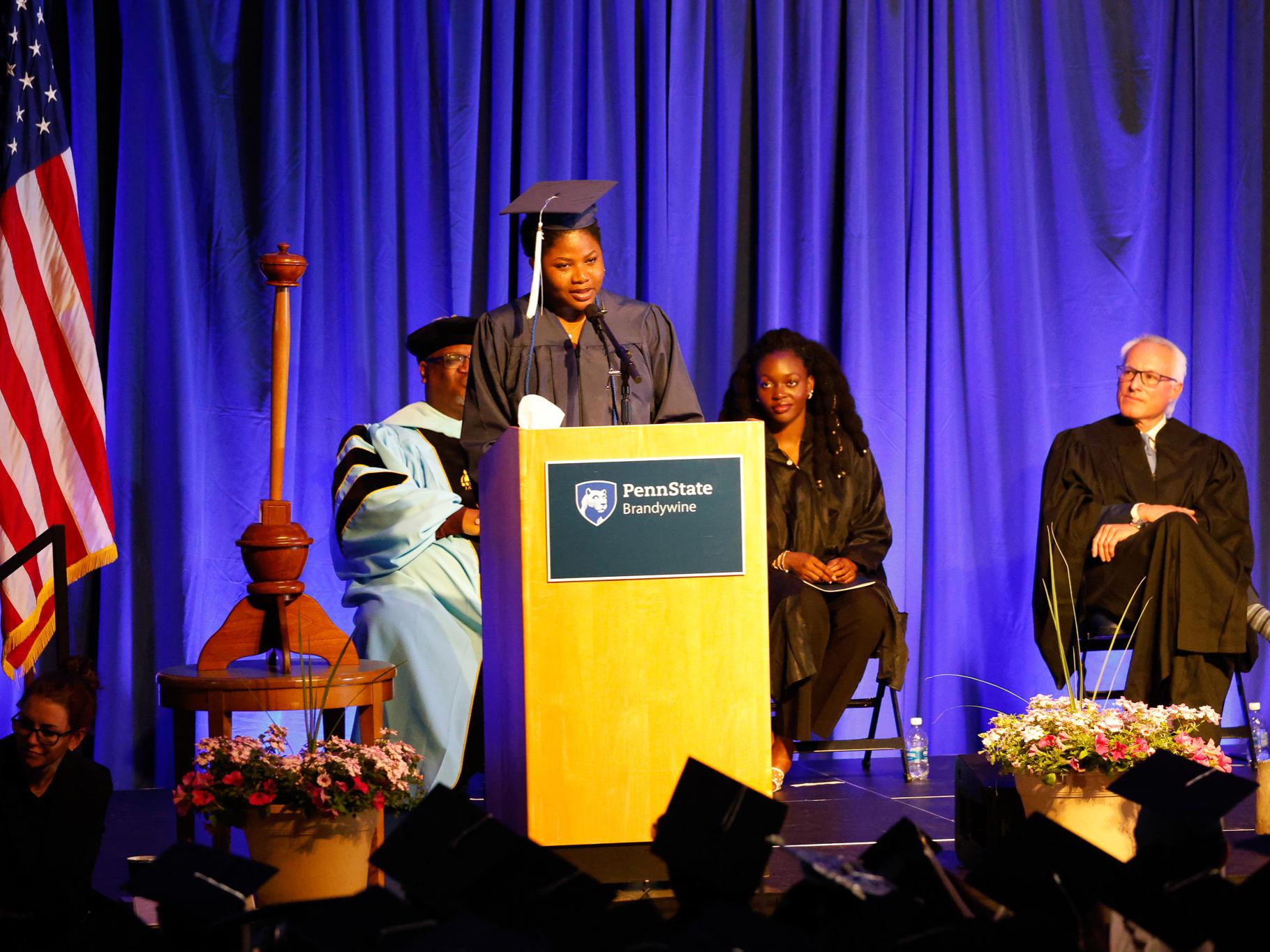 Catherine Adewumi speaking at a podium wearing a cap and gown with Austin Duckett, Jemimah Mwaba and Joseph Kernen sitting behind her on stage