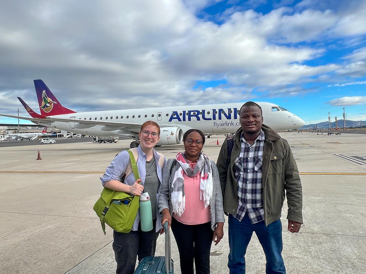 Isabel, Ida, and Abdul arrive in South Africa, standing on an airport tarmac in front of an Airlink airplane with luggage and travel bags under a partly cloudy sky.