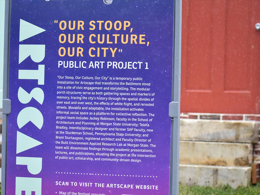 Signboard for a public art project titled “Our Stoop, Our Culture, Our City,” with descriptive text about a temporary Baltimore installation and a red door facade visible to the side.