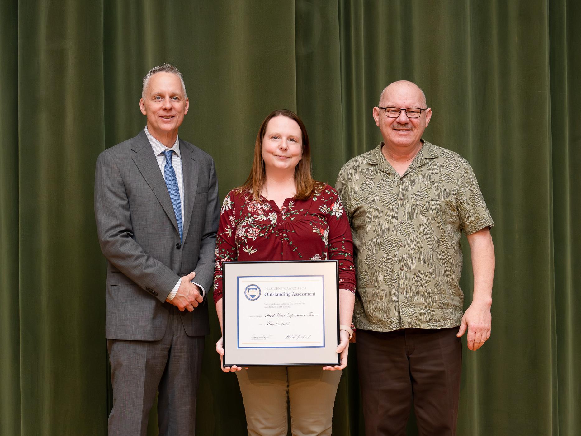 Three people posing for a photograph, the one in the center holds up an award