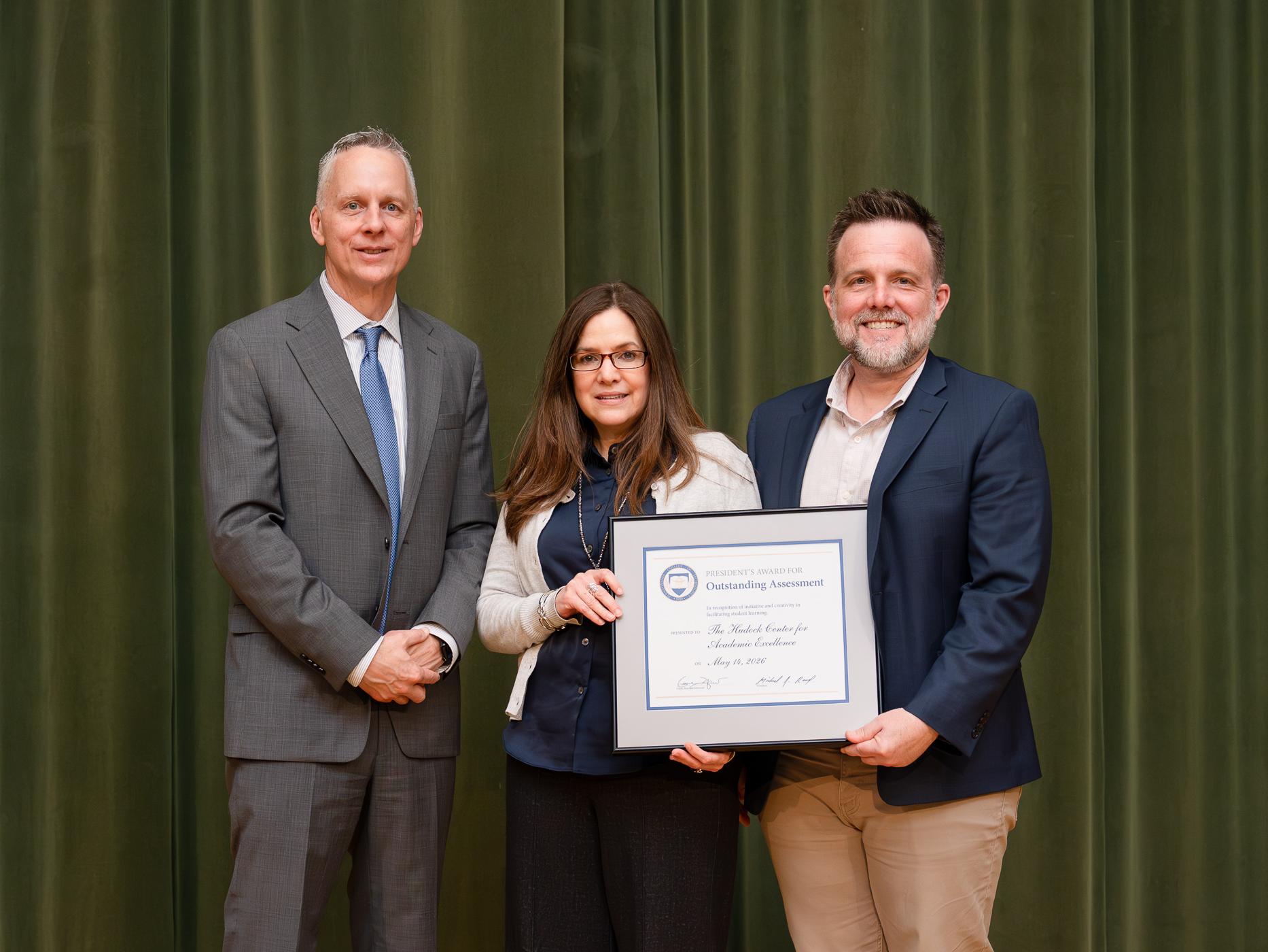 three people pose for a photo, the one in the center is holding up an award