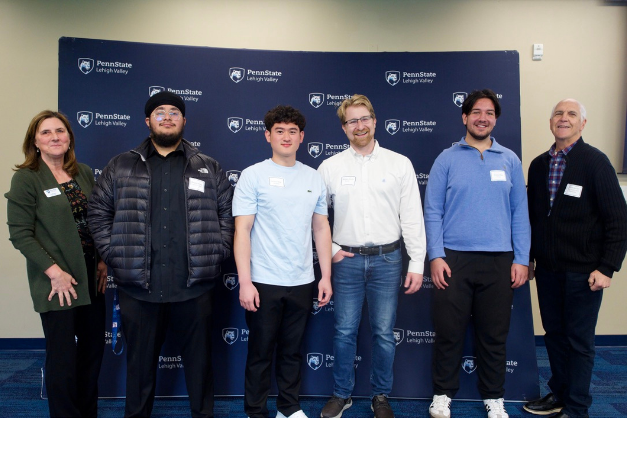 a male professor stands next to his students smiling in front of a step and repeat banner