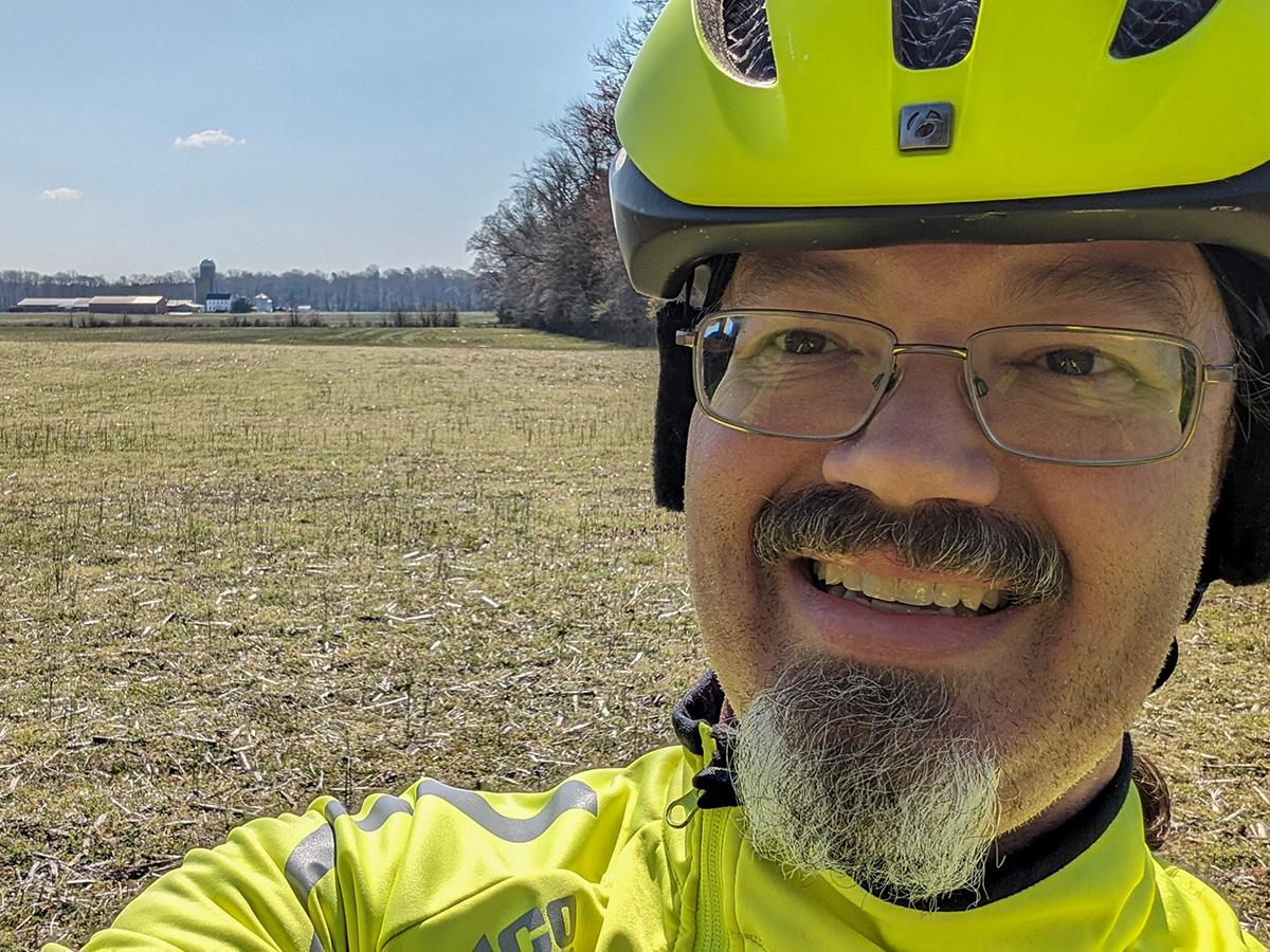 Kroon wears a bright yellow cycling helmet and jacket smiles in a sunny field. Behind him, trees and a distant barn are visible under a clear blue sky.