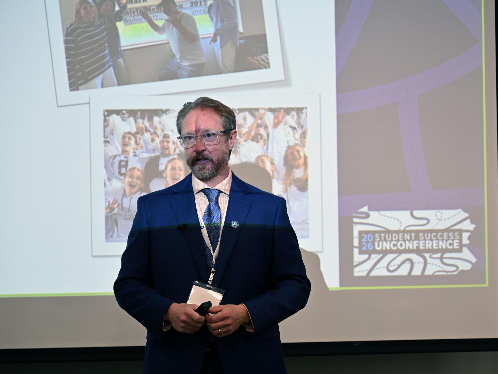 A man wearing a blue suit stands at the front of a room and speaks.